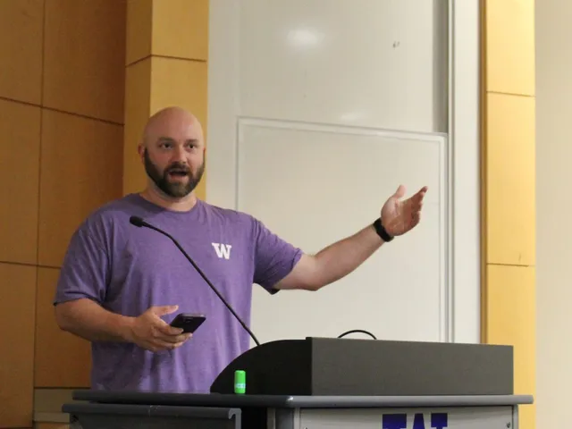 A man in a purple University of Washington T-shirt speaking and gesturing at a podium bearing a large block W logo.