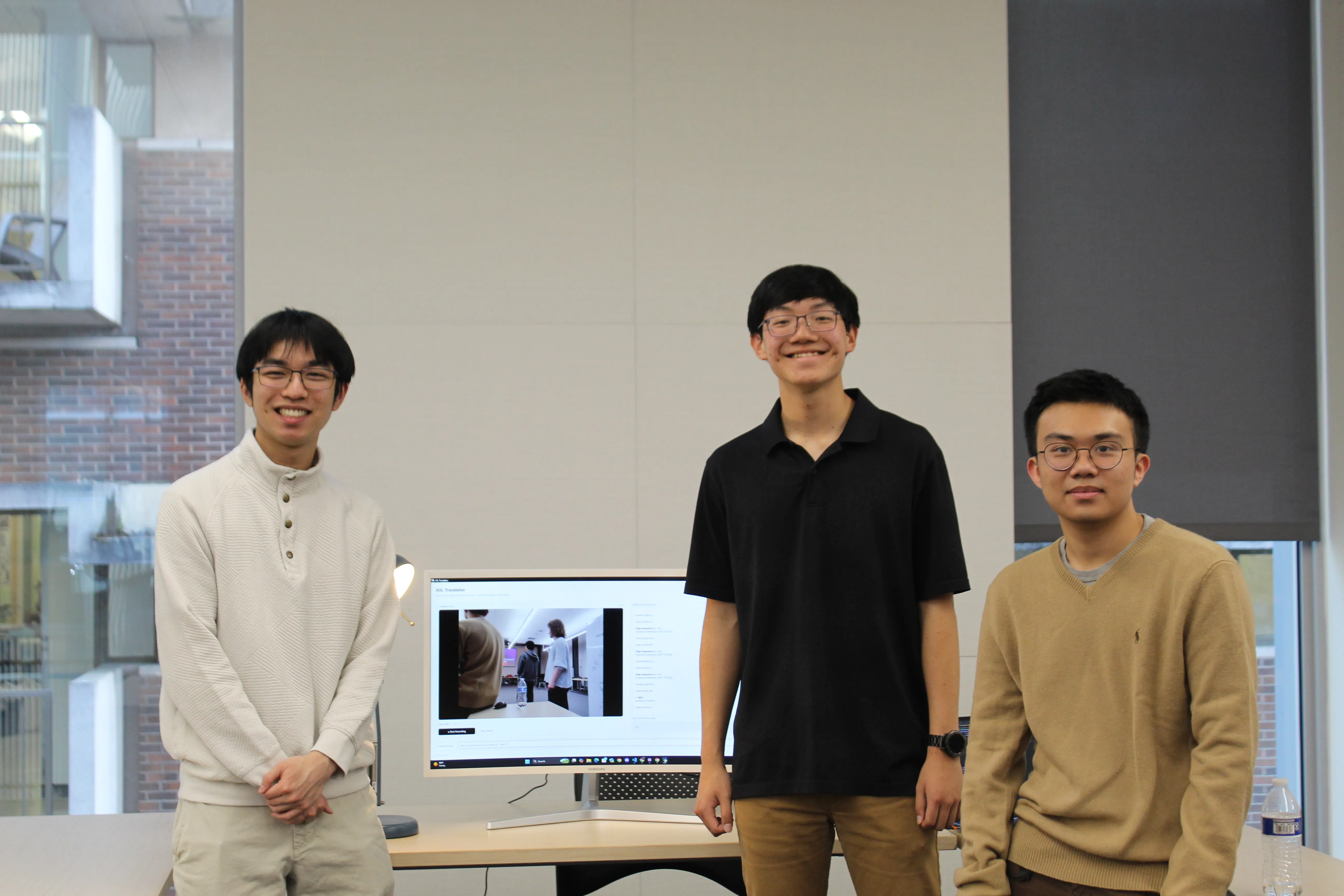 Three students smiling indoors next to a monitor displaying their ASL Translator app, showing a live camera view of a person signing.