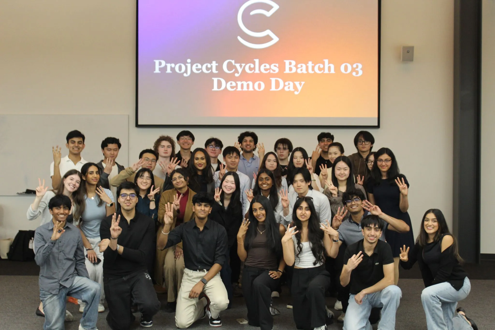 A large group of students posing together and holding up three fingers, in front of a screen reading "Project Cycles Batch 03 Demo Day."