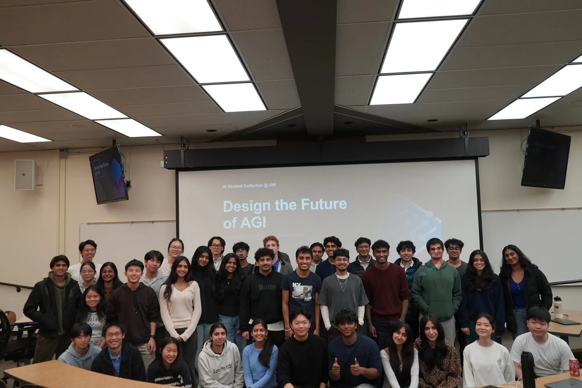 About fifty students posing together at the front of a lecture hall, under a slide reading "Design the Future of AGI."
