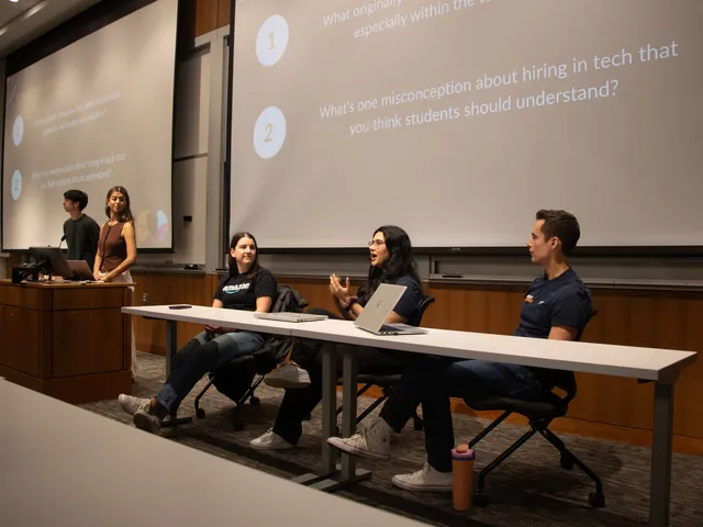 Three recruiters, one in an Amazon T-shirt, seated behind a table on a lecture-hall stage, the one in the middle gesturing as she speaks, under a slide asking about misconceptions in tech hiring; two student hosts stand at the podium.