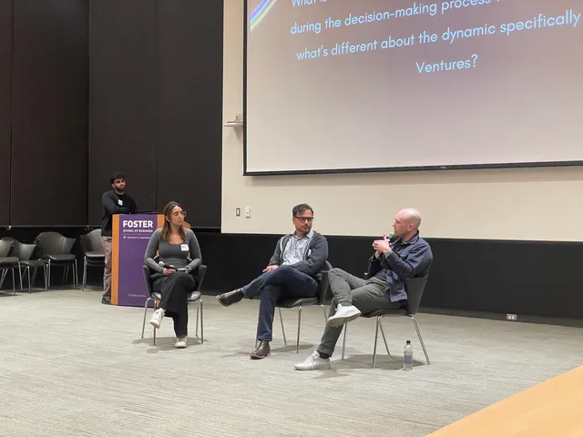 Two investors and a student moderator seated on a stage at the Foster School of Business, one investor gesturing with a microphone as he answers a question about Pack Ventures projected on the screen behind them.