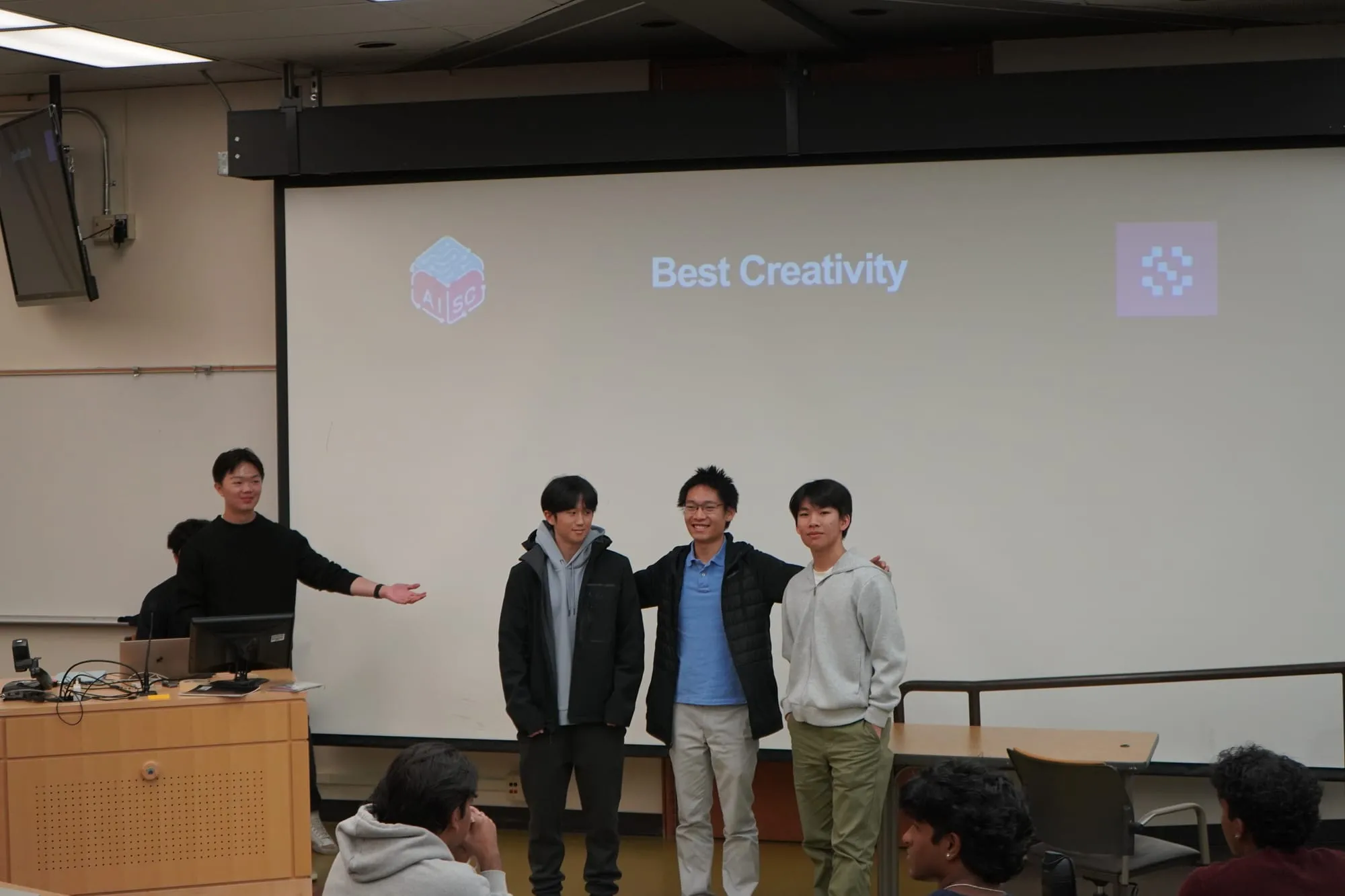 Three students standing in front of a screen reading "Best Creativity" as an organizer gestures toward them.