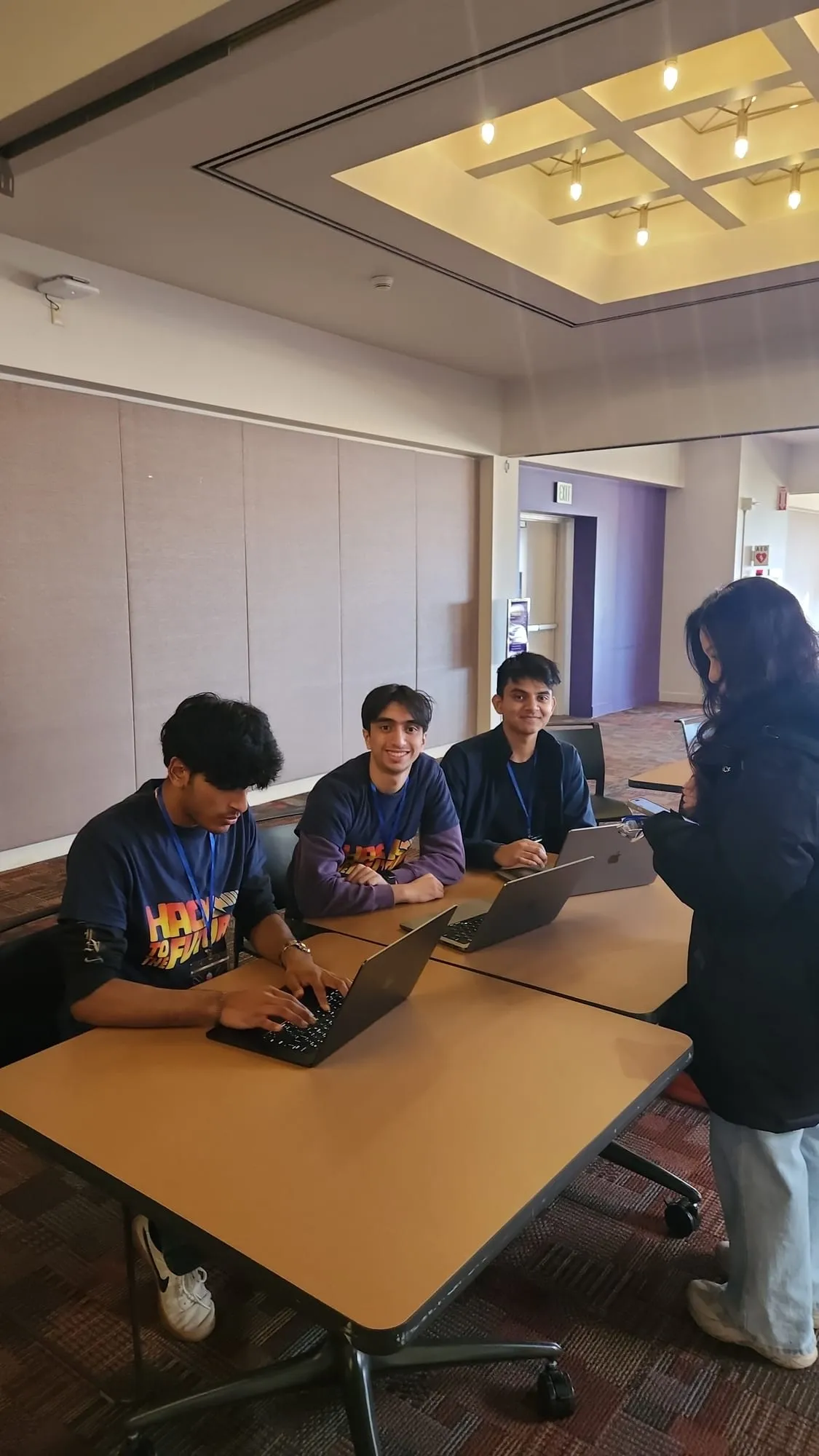 Three students in hackathon T-shirts working on laptops at a table while a fourth person stands beside them.
