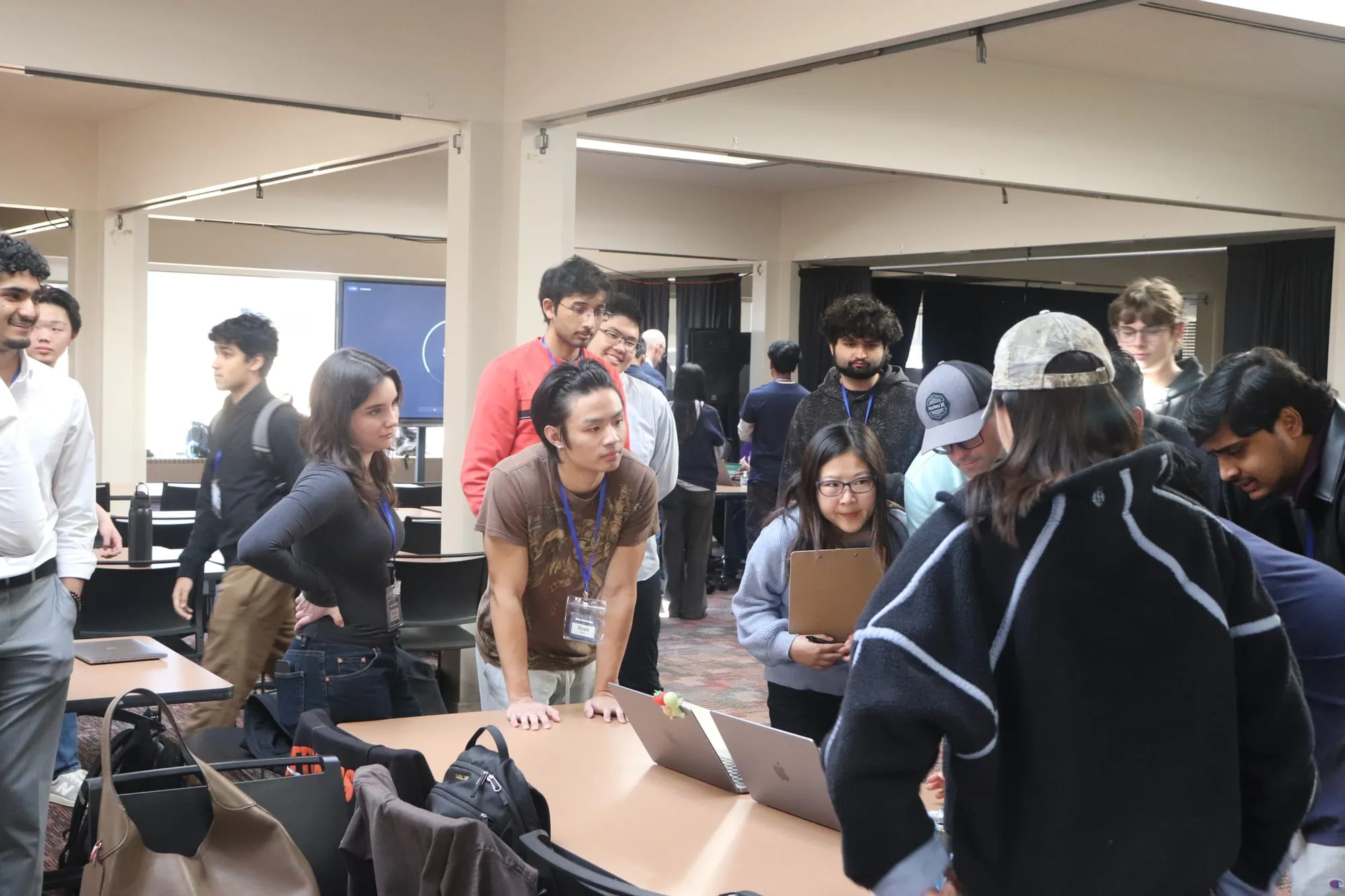 Students and judges crowding round a table of open laptops during hackathon demos.