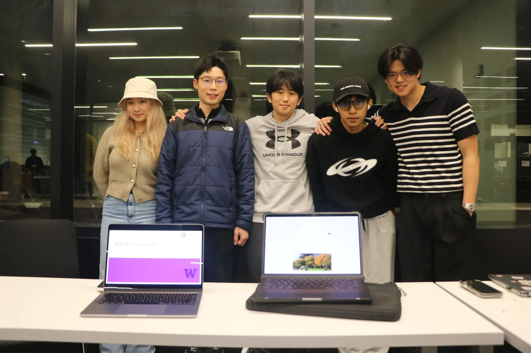 Five students standing behind a table with two open laptops, one displaying the HuskyConnect app's title screen with a University of Washington W logo.