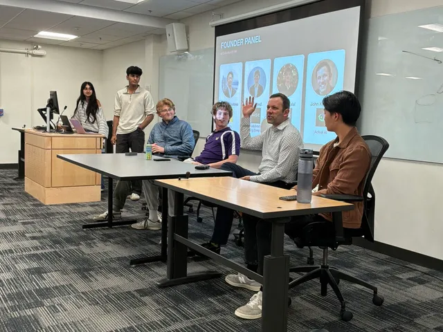 Four panelists and two students seated and standing around tables, one panelist gesturing while speaking, in front of a projected "Founder Panel" slide showing headshots of four speakers.